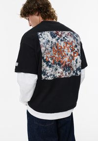 Ung mand med krøllet hår iført lagdelte sorte og hvide langærmede skjorter samt en sort T-shirt med et abstrakt mønster i rød og blå på ryggen.