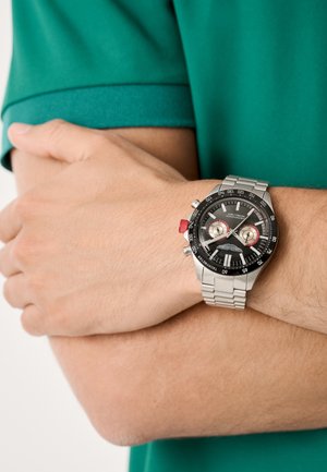 Orologio da polso in acciaio inossidabile con quadrante nero, rifiniture rosse e funzionalità cronografo. Indossato su una mano contro un braccio, con una maglietta polo verde.