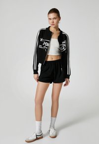 Sort cropped bomberjakke med hvide striber og grafiske prints, parret med sorte shorts og hvide sneakers, hvilket skaber et sporty sæt.