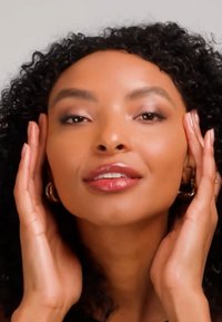 Femme aux cheveux bouclés touchant doucement son visage, portant des boucles d'oreilles anneaux et un maquillage naturel, sur un fond clair uni.