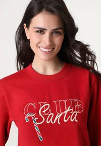 Felpa rossa con la scritta "CLUB Santa" e un dettaglio a bastoncino di zucchero. Realizzata in tessuto morbido, presenta uno scollo rotondo e una vestibilità comoda.