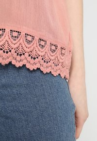 Blusa rosa con un bordo in pizzo all'orlo, caratterizzata da bordi ondulati e motivi floreali. Indossata sopra dei jeans di denim blu. Tessuto con texture.