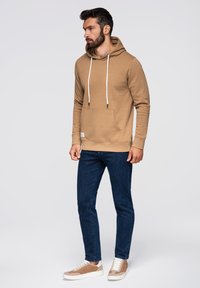 Hnědá mikina s kapucí, přední kapsou, bílými šňůrkami a manžetami, spárovaná s tmavě modrými slim-fit džíny a světle hnědými teniskami.