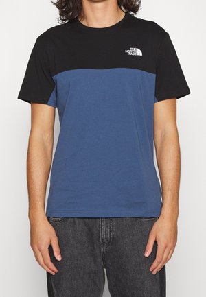 Osoba nosząca czarno-niebieską koszulkę z krótkim rękawem, z blokowym wzorem i logo The North Face na górnej lewej części klatki piersiowej.