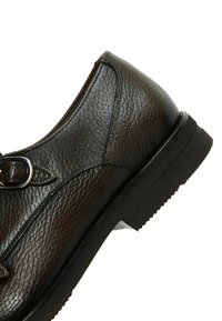 Musta nahkakenkä, jossa on teksturoitu pinta, solkisomiste ja litteä pohja. Malli on pyöreä, ja siinä on hillittyjä ompeluaksentteja.