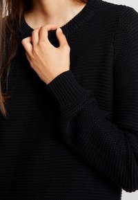 Maglione nero lavorato a maglia con scollatura rotonda e polsini a costine. Presenta un tessuto texture e un primo piano di una mano vicino al colletto.