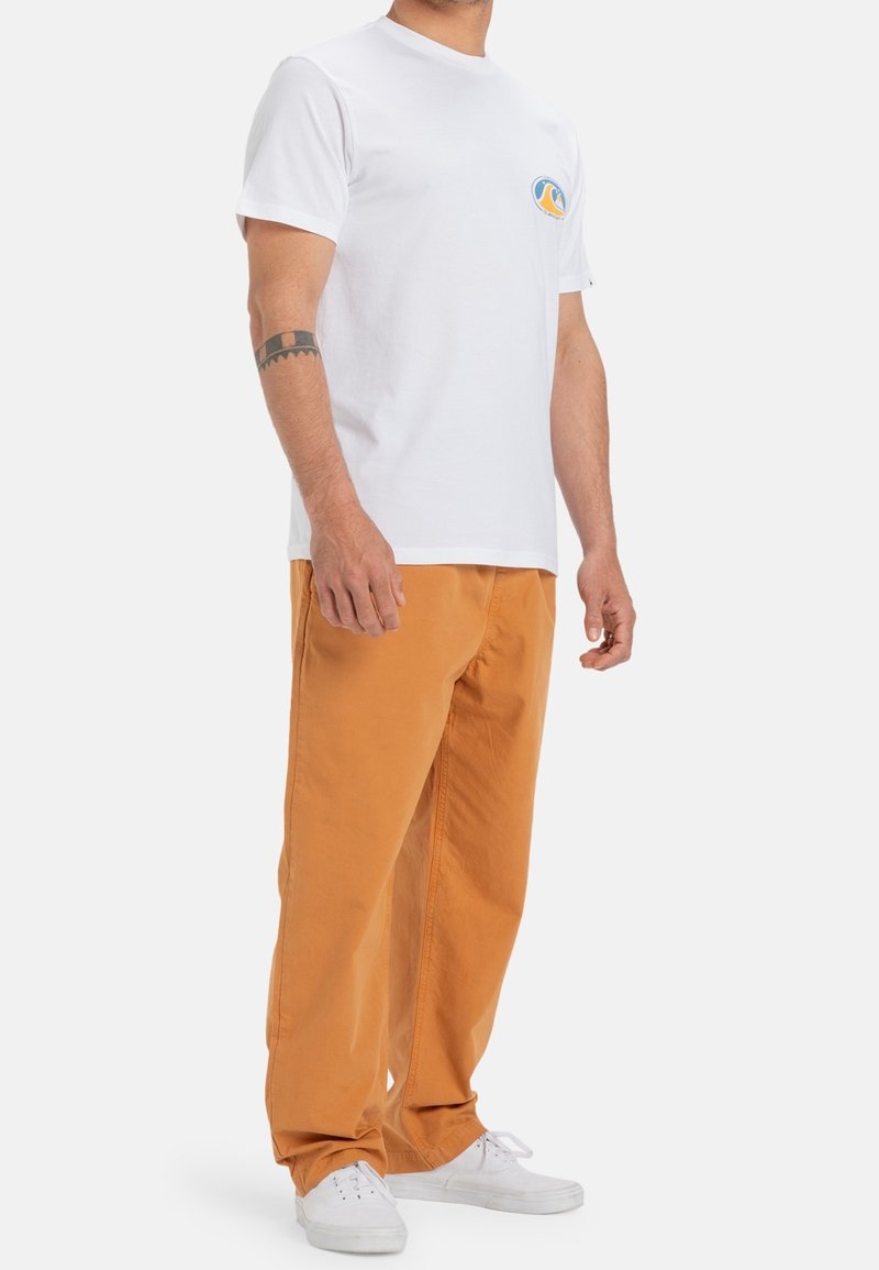 Homme portant un t-shirt blanc avec un petit logo, un pantalon orange ample, des baskets blanches et un tatouage au poignet, debout devant un fond uni.