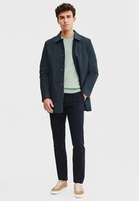 Ciemnoniebieski płaszcz, jasnooliwkowy sweter, granatowe spodnie i beżowe trampki. Gładki materiał z zapięciem na guziki; buty mają sznurowadła i kontrastującą podeszwę.