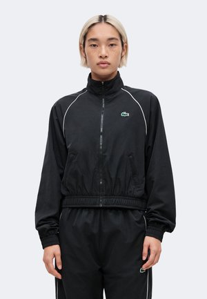 Persoon met kort blond haar draagt een zwarte zip-up jas en bijpassende broek met witte biezen en Lacoste-logo's op borst en dij.