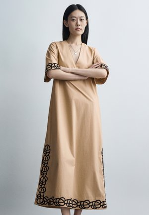 Femme debout, bras croisés, portant une robe beige avec des motifs circulaires noirs sur les manches et l'ourlet, sur un fond gris uni.