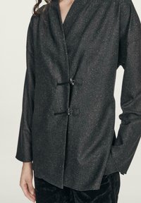 Sort struktureret jakke med et omslagsdesign, to hægter, lange ærmer og en subtil glans. Blødt stof med spredte detaljer.