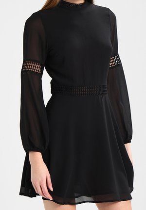 Sort kjole med gennemsigtige lange ærmer, hæklede detaljer ved taljen og på de øverste arme samt en høj halsudskæring.