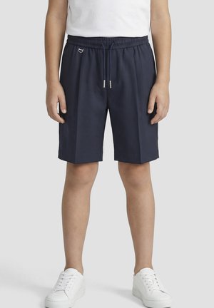 Persona vistiendo pantalones cortos azul marino hasta la rodilla con cordón y zapatillas blancas, de pie contra un fondo claro y liso.