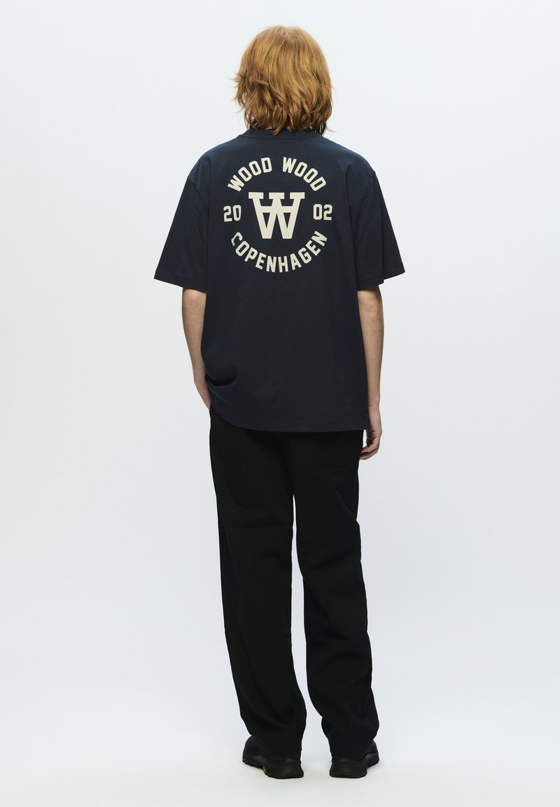 Marineblå bomulds t-shirt med grafikken "WOOD WOOD COPENHAGEN 2002" i cremefarve, med en løs pasform og korte ærmer, parret med sorte bukser.