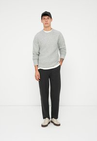 Lys grå strikkegenser med teksturert mønster, lagd over en hvit skjorte. Mørke, løse bukser og kremfargede sneakers med brune såler. Svart caps.