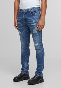 Jeans de mezclilla azul desgastada con detalles desgarrados y remendados, de corte ajustado y sutil salpicadura de pintura. Combinados con zapatos de cuero negros.