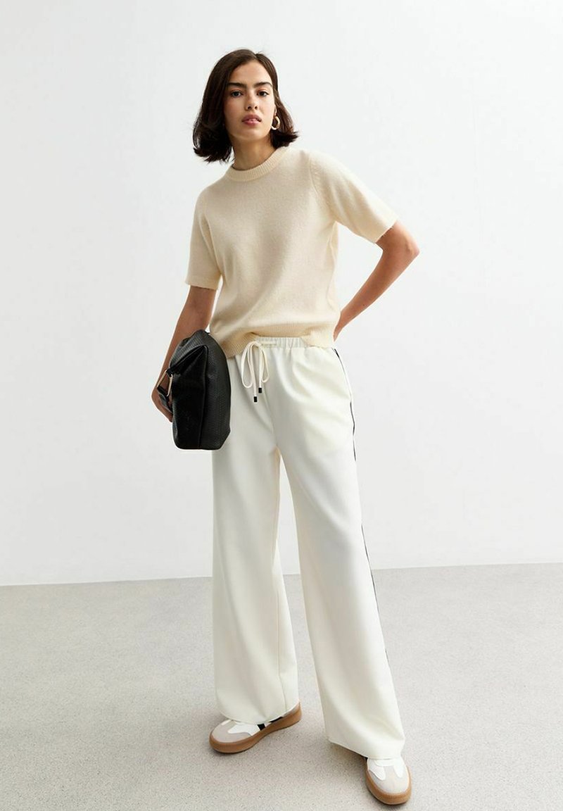 Un pull Beige clair à manches courtes associé à un pantalon blanc avec cordon de serrage et bandes latérales. Le mannequin tient un sac noir texturé. Chaussures : baskets beiges.