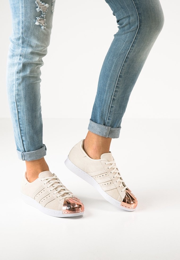 Beige mockaskor med en glansig roséguld tåhätta, vit gummisula och en minimalistisk snörningsdesign. Slitna blå jeans är på.