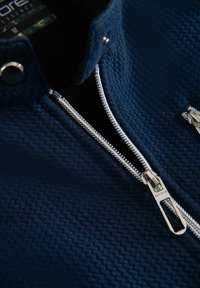 Marinblå jacka med textur och hög krage, som har en silverfärgad dragkedja och tryckknappsstängning. Tyget har ett vävt mönster.