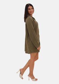 Olivengrøn skjortekjole med tekstureret stof, knaplukning foran, lange ærmer og løs pasform; kombineret med hvide åbne hæle-sandaler.