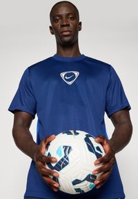 Μπλε αθλητικό μπλουζάκι με στρογγυλή λαιμόκοψη και λευκό λογότυπο Nike, το οποίο φοράει ένα άτομο που κρατά μια πολύχρωμη μπάλα ποδοσφαίρου με μπλε και πορτοκαλί λεπτομέρειες.