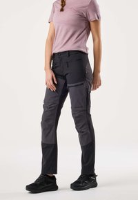 Frau trägt schwarze und graue Cargo-Hose mit mehreren Reißverschlusstaschen und schwarze Wanderschuhe, steht vor weißem Hintergrund.