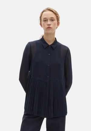 Camicia blu navy con apertura a bottoni, maniche trasparenti e dettagli plissettati nel torso. Tessuto morbido con una silhouette strutturata.