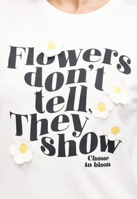 Λευκό πουκάμισο με μαύρο κείμενο "Τα λουλούδια δεν λένε, δείχνουν" και "Επέλεξε να ανθίσεις," διακοσμημένο με πέντε λευκά και κίτρινα πλεκτά λουλούδια.