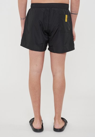 Pantaloni da bagno neri con una cintura elastica, una tasca posteriore con un'etichetta del marchio gialla e una texture leggera e liscia.