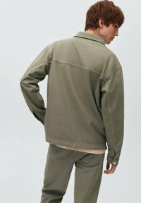 Olivnozeleni denim jakna s ovratnikom, dolgimi rokavi in zadnjim šivom yoke, v kombinaciji z usklajenimi hlačami. Ima sproščen kroj in vidno šivanje.