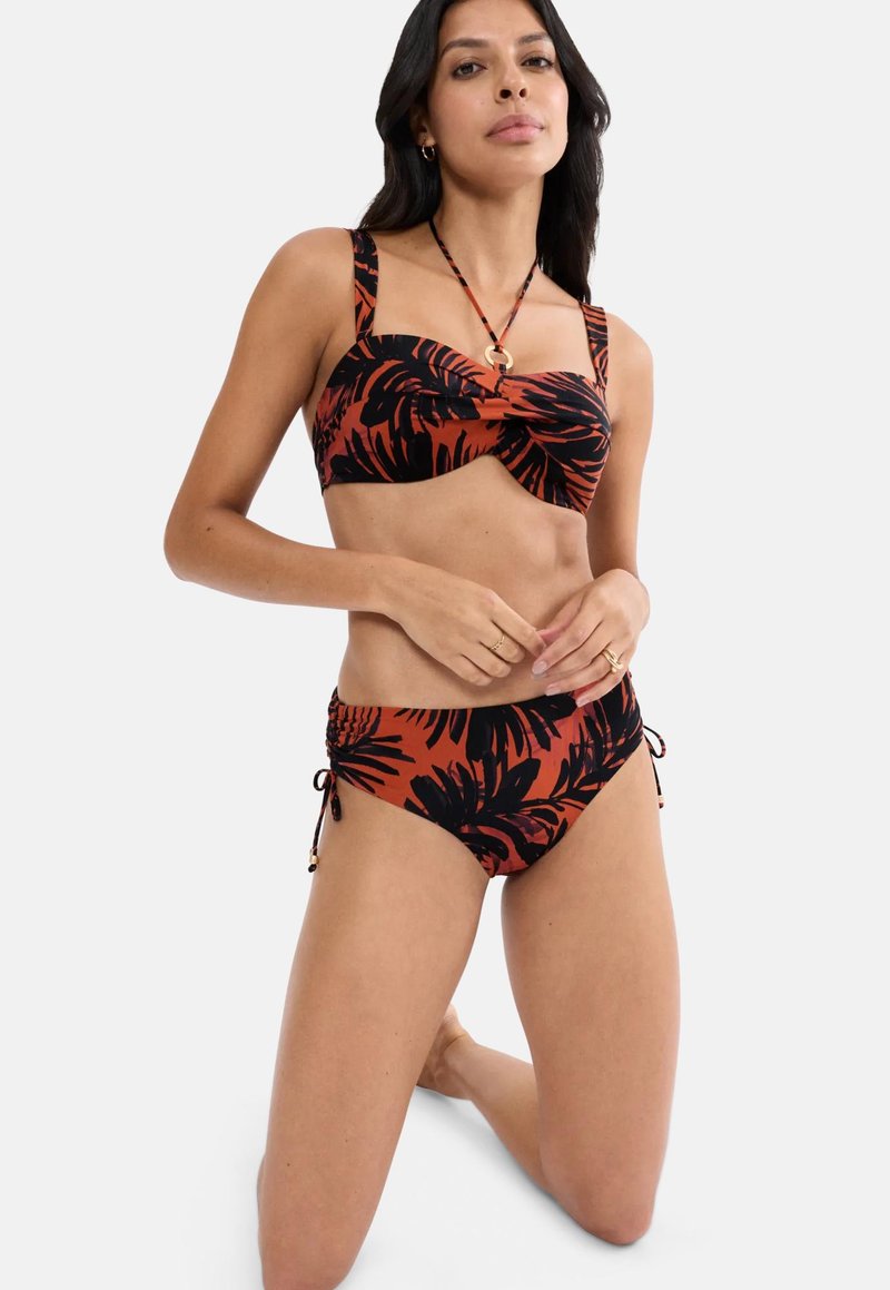 Femme à genoux portant un bikini à imprimé tropical rouille et noir, avec un haut bandeau torsadé et un bas taille haute avec des liens sur les côtés.