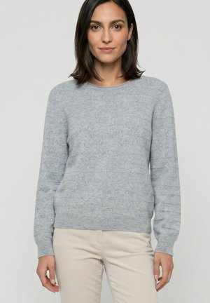 Vrouw met steil donker haar, die een lichtgrijze gestructureerde trui en beige broek draagt, staand tegen een effen achtergrond.