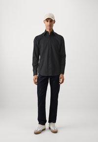Schwarzes Langarmhemd mit einem button-down Kragen, kombiniert mit dunklen Jeans und weißen Sneakern mit beigen Akzenten und Schnürsenkeln.