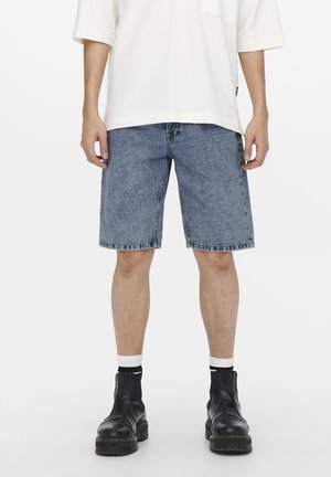 Personne portant un short en denim bleu clair, des chaussettes blanches mi-hautes, des bottes en cuir noir et une chemise blanche à manches courtes ample, debout devant un fond blanc.