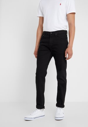 Person iført cuffede sorte jeans, hvid polo med rødt logo og hvide sneakers, stående mod en ensfarvet lys baggrund.
