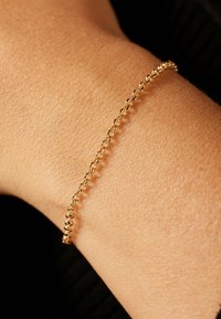Delicate gouden kettingarmband met ovale schakels, gedragen om een pols tegen een donkere stof achtergrond.