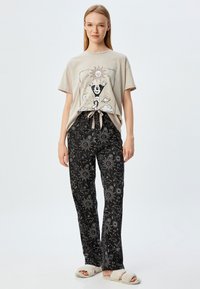 Beige graphic T-shirt met een knoop aan de voorkant, gecombineerd met zwarte broeken met een zon- en wolkenpatroon. Gezellige instap slippers maken de look af.