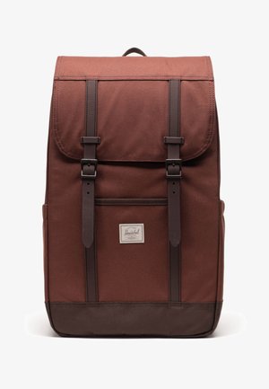 Mochila de tela marrón con solapa superior, acentos de cuero y dos correas ajustables. Cuenta con un bolsillo frontal y un parche con logotipo.