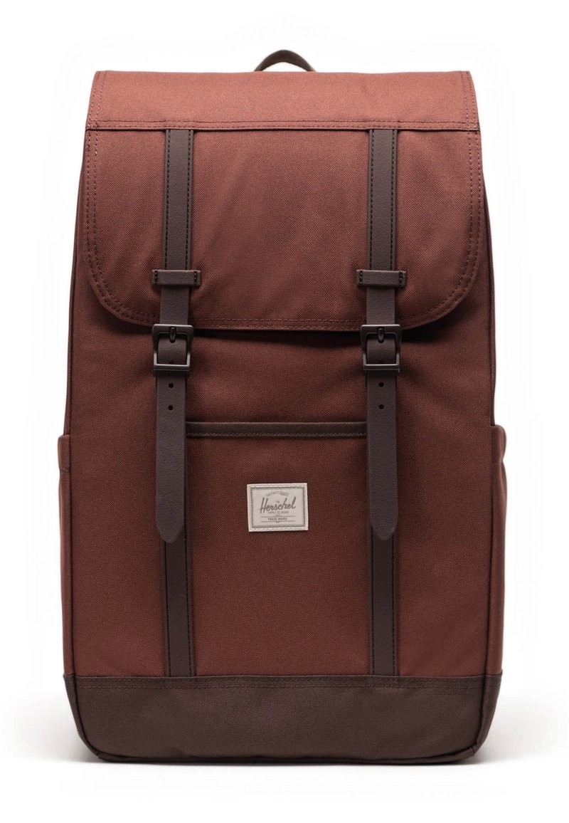 Mochila de tela marrón con solapa superior, acentos de cuero y dos correas ajustables. Cuenta con un bolsillo frontal y un parche con logotipo.