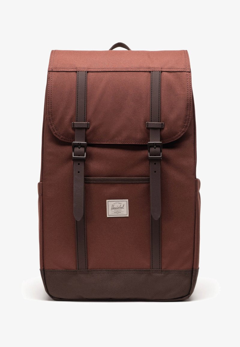 Mochila de tela marrón con solapa superior, acentos de cuero y dos correas ajustables. Cuenta con un bolsillo frontal y un parche con logotipo.