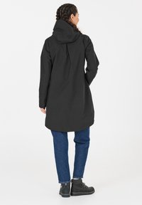 Svart vattentät jacka med huva, har en rundad fåll och en avslappnad passform. Bärs över blå jeans och svarta stövlar.