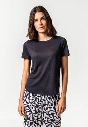 Frau mit schulterlangem braunem Haar, die ein schlichtes dunkles Kurzarmshirt und gemusterte schwarz-weiße weite Hosen trägt, steht vor weißem Hintergrund.