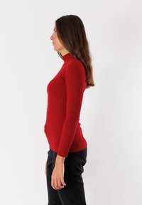 Maglione rosso a collo alto con coste, maniche lunghe, design aderente, tessuto testurizzato e un collo alto. Indossato con pantaloni neri in vista laterale.