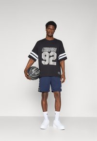 Fekete, oversized póló "CHAMPION 92" felirattal és fehér csíkokkal, tengerészkék sorttal, amelyen logós szalag található, fehér sportcipővel és kosárlabdával párosítva.