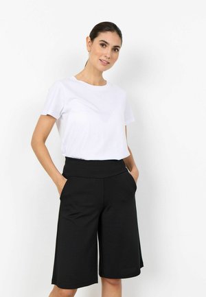 Kvinde stående med hænderne i lommerne iført en ensfarvet hvid t-shirt og sorte, vidde knælange shorts mod en hvid baggrund.