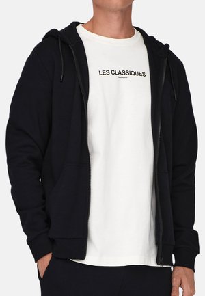 Schwarze Zip-Hoodie mit verstellbarem Kordelzug, zwei frontalen Taschen und gerippten Bündchen, getragen über einem weißen T-Shirt mit schwarzem Text.