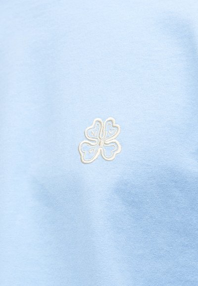 Ανοιχτό μπλε ύφασμα με μια μικρή λευκή κεντημένη τετράφυλλη τριφύλλι στο κέντρο της υφασμάτινης επιφάνειας.