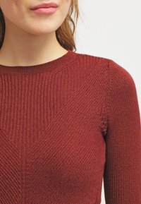 Ribbad vinröd tröja med rund halsringning, texturerad tyg och diagonala stickade mönster, vilket visar en figursydd och strukturerad design.
