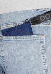 Navy fabric wallet partially visible in a denim pocket, featuring a geometric pattern, blue stitching, and an attached black strap.