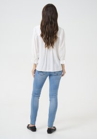 White long-sleeve blouse with gathered sleeves, paired with light blue skinny jeans and black flat shoes, viewed from the back.